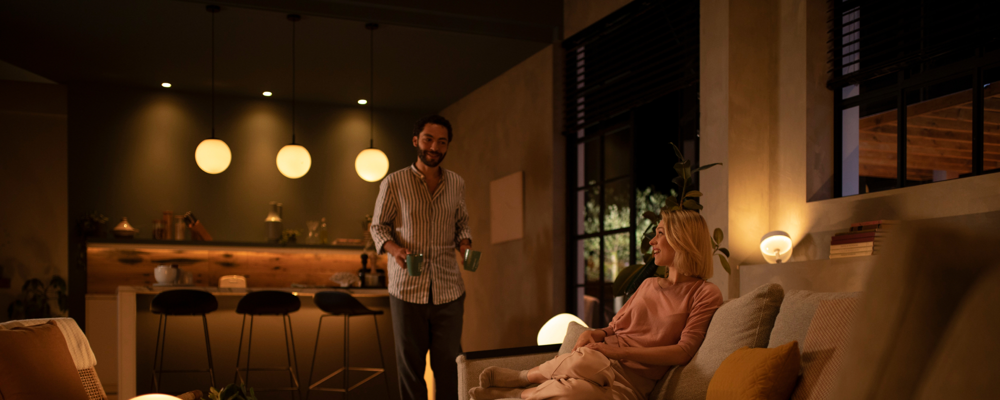 Ein Paar entspannt sich zufrieden im Wohnzimmer, das von smartem Licht in warme Weißtöne gehüllt ist, um den Philips Hue Neujahrsverkauf anzukündigen.